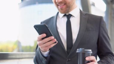 A young confident businessman with a coffee cup using a mobile phone near a business center