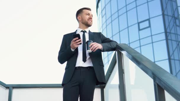 A well dressed happy bearded office corporate male executive standing outside a modern corporate building with coffee cup and cellphone — Stockvideo