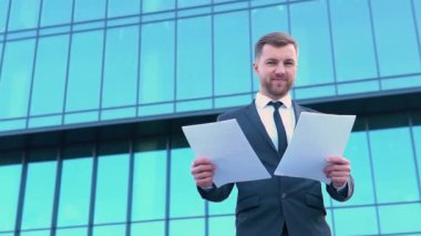 Modern bir ofis binasının önündeki belgelerle takım elbiseli özgüvenli bir adamın portresi.