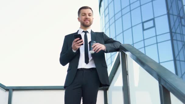 Businessman standing outside a modern corporate building with coffee cup and cellphone. Cityscape background — Vídeos de Stock