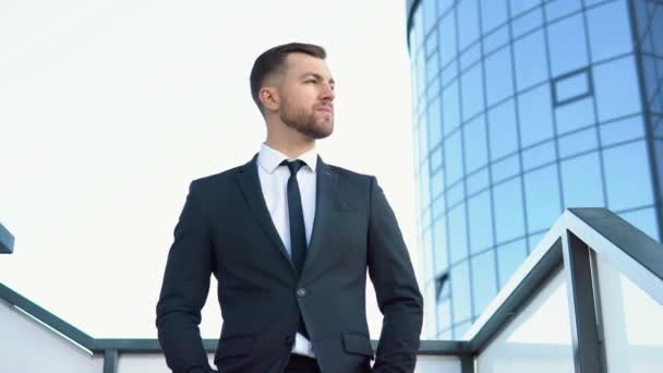 Un homme de bureau barbu heureux bien habillé dirigeant d'entreprise debout à l'extérieur d'un bâtiment d'entreprise moderne et regardant dans la caméra — Video