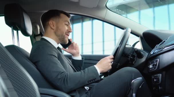 A confident man speaks on the phone sitting in a car in a business suit near a modern office building — Stockvideo