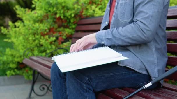 Слепой читает книгу Брайля, сидит на скамейке в летнем парке — стоковое видео
