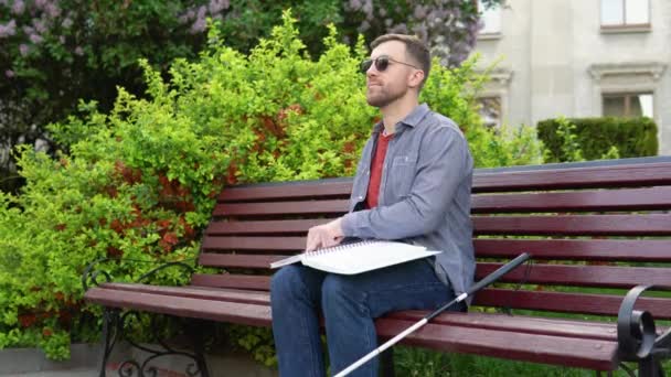 Fiatal vak férfi ül a padon a Városligetben, és Braille könyvet olvas. — Stock videók