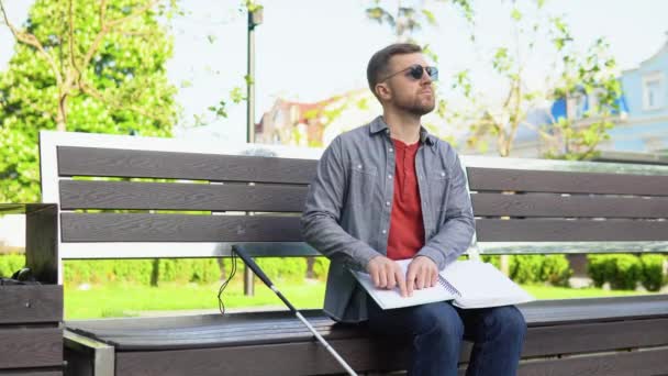 Joven ciego sentado en el banco en el parque de la ciudad y leyendo un libro Braille — Vídeo de stock