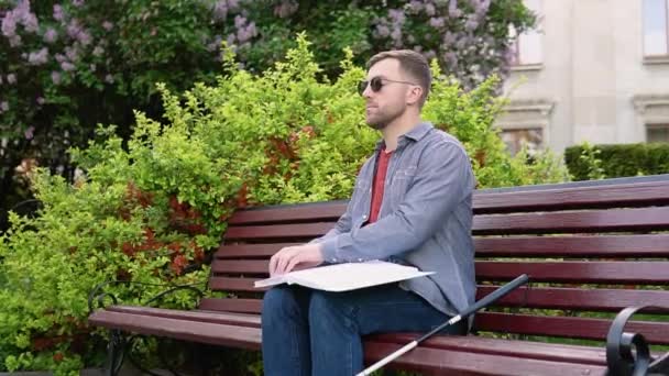Młody niewidomy siedzi na ławce w parku i czyta książkę Braille 'a — Wideo stockowe