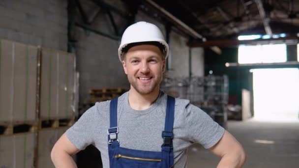 잘 생기고 행복 한 직업적 근로자, 모자를 쓰고 웃으면서 카메라에 미소짓는 사람. 뒤에 있는 큰 창고에서 — 비디오