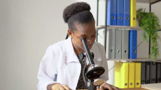 顕微鏡でウイルスを観察するアフリカ系アメリカ人女性医師。女性科学者は研究室で顕微鏡を通して研究をしています — ストック動画