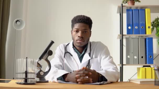 Африканский американский врач смотрит в камеру, сидя за столом в больнице — стоковое видео