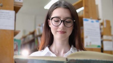 A thoughtful young student reads a book standing in the library. A smiling young girl in glasses reads a book in the university library
