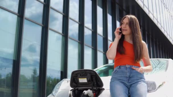 ショッピングセンターの近くで電気自動車を充電しながら電話で話す魅力的な女性 — ストック動画
