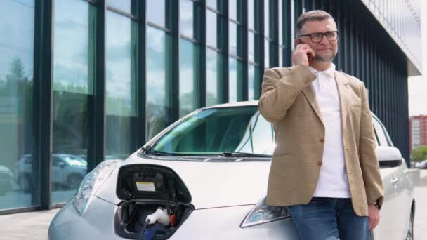 Senior Man Speaks Phone While Electric Car Charging Outdoors — Stock Video