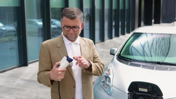 老人は電気自動車の近くで充電器を持って立っている 充電ステーションから電気自動車を充電するプラグ — ストック動画