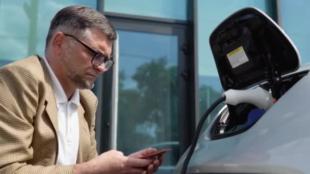 Starszy Mężczyzna Odłącza Kabel Ładowania Pojazdu Elektrycznego Używa Telefonu Odłączenia — Wideo stockowe