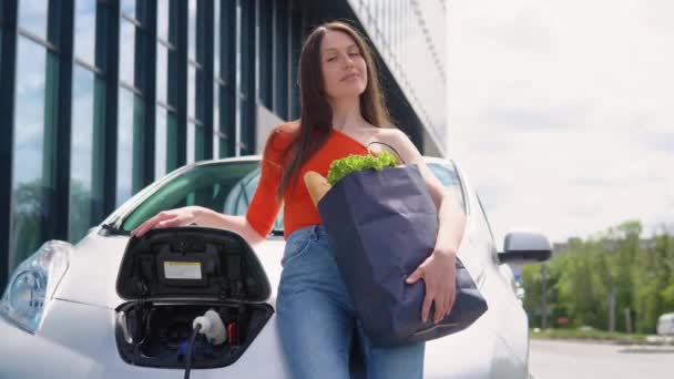 女性は食料品と車のそばに立っている 電気ガソリンスタンドでの電気自動車の充電 — ストック動画