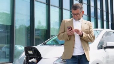 Üst düzey işadamı akıllı telefon kullanıyor ve elektrikli araçlara güç kaynağı bağlayıp arabadaki aküyü şarj ediyor. Şarj istasyonundan bir elektrikli arabayı şarj etmek