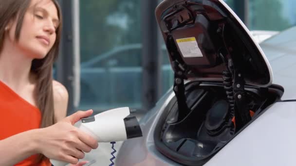 女性は電気自動車に充電器を入れて待っているときに電話を使う 環境に配慮した技術 再生可能エネルギーの概念 エミッション電気自動車に接続された充電コード — ストック動画