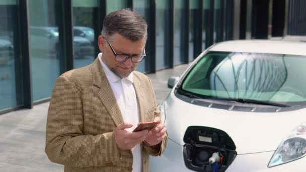 Elektrikli Arabayı Şarj Ederken Akıllı Telefon Kullanan Yakışıklı Bir Adamı — Stok video
