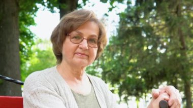 Şehir parkındaki bankta gülümseyen gözlüklü yaşlı kadın portresi. Mutlu beyaz kadın kameraya bakıyor.