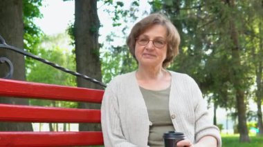 Şehir parkındaki bankta gülümseyen gözlüklü yaşlı kadın portresi. Mutlu beyaz kadın kameraya bakıyor.