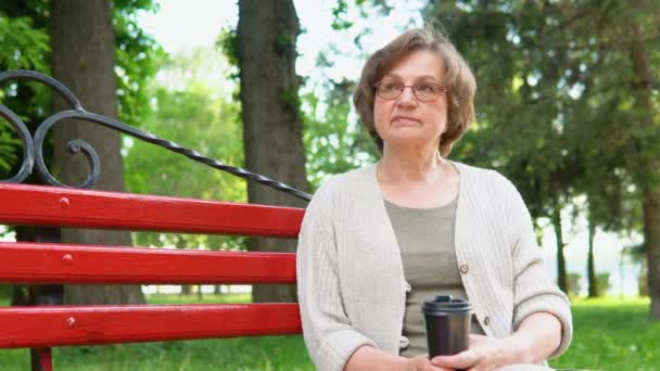 ベンチの上の公園に座っているスマートフォンとコーヒーを持つ高齢女性の肖像 — ストック動画