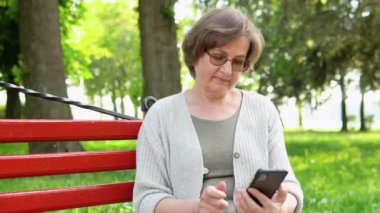 Parktaki bankta oturan yaşlı kadın telefonla konuşuyor.