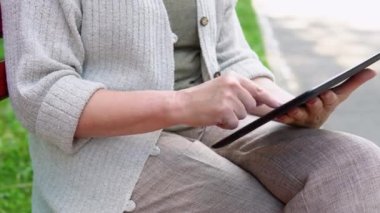 Elinde akıllı telefon olan yaşlı bir kadın bir bankta oturuyor, internette geziniyor ve sosyal ağlarda haberleri araştırıyor.