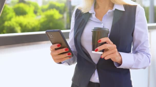 コーヒーカップと携帯電話を備えたモダンな企業ビルの外に立っているシニアビジネス女性 都市景観 — ストック動画