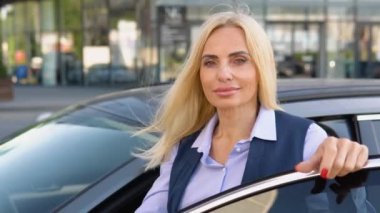 İş elbisesiyle arabasının yanında duran kendinden emin yaşlı bir kadının portresi bir iş merkezinin arka planında duruyor.
