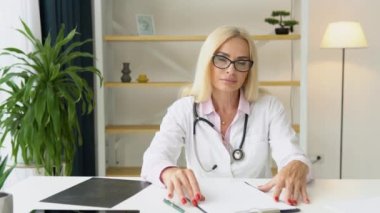 Beyaz ceketli ve steteskoplu yaşlı Avrupalı kadın doktor kameraya bakıyor.