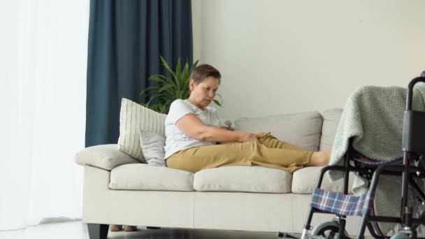 Mulher Idosa Massageando Pernas Enquanto Senta Sofá Perto Cadeira Rodas — Vídeo de Stock