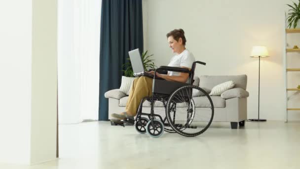坐在轮椅上的成熟女人笑着工作 一个人在家用笔记本电脑 网上学习或购物 — 图库视频影像