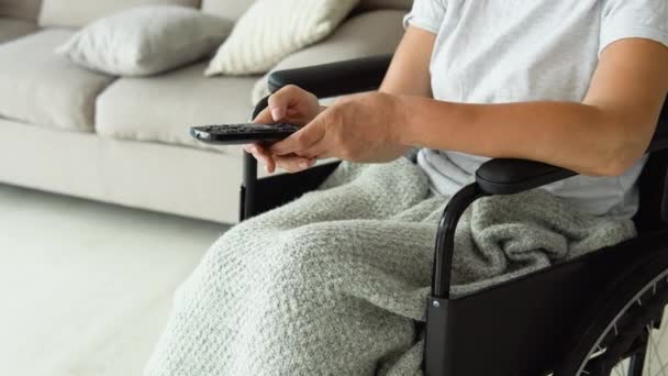 テレビチャンネルを変更するリモートコントロールを保持し リビングルームでテレビを見て車椅子のシニア女性 おばあちゃんはテレビをつけている 自宅でのレジャー — ストック動画