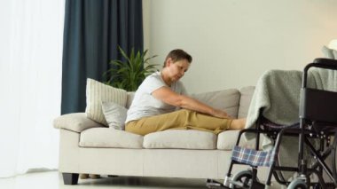 Yaşlı bir kadın tekerlekli sandalyenin yanındaki koltukta otururken bacaklarına masaj yapıyor.