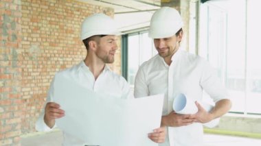 A young engineers in safety helmets standing on an under-construction building holding a blueprint of the structure and working together