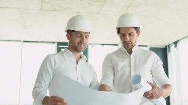 A young engineers in safety helmets standing on an under-construction building holding a blueprint of the structure and working together