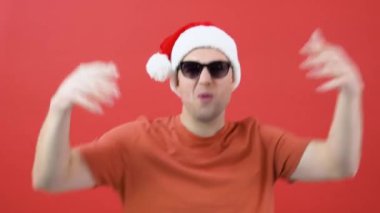 Turuncu tişörtlü, beyaz, orta yaşlı bir adam güneş gözlüğü takıyor ve Noel Baba şapkası kollarını sallıyor. Kırmızı Noel kıyafetli adam kırmızı bir arka planın önünde duruyor.