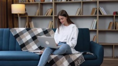 Evdeki oturma odasında kanepede otururken gümüş dizüstü bilgisayar kullanan güzel, neşeli kız..