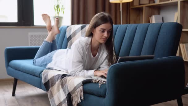 リラックスした笑顔でインターネットをサーフィンするラップトップ コンピューターを使用してソファ彼女の胃に横になっている若い女性 — ストック動画