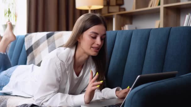 年轻女子躺在她的肚子上沙发上使用手提电脑上网与快乐轻松的微笑 — 图库视频影像