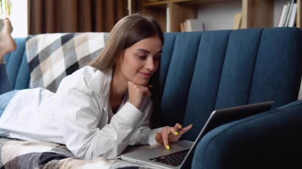 リラックスした笑顔でインターネットをサーフィンするラップトップ コンピューターを使用してソファ彼女の胃に横になっている若い女性 — ストック動画