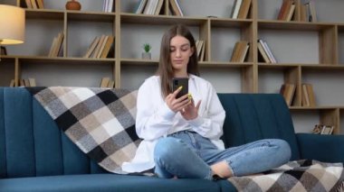 Happy woman using mobile phone while sitting a couch at home with laptop computer