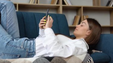 Gülümseyen güzel kız evde bir kanepede rahatlatıcı, cep telefonu kullanarak