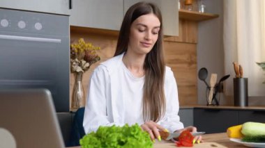 Yemek blogcusu kavramı. Mutfak bilgisayarında yemek pişirmekten zevk alan genç ve mutlu bir kadın. Mutfak masasındaki dizüstü bilgisayarda yemek tarifi arıyor..