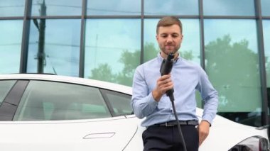 Yakışıklı adam yeni elektrikli arabasının yanında duruyor ve şarj aletinin fişini tutuyor..