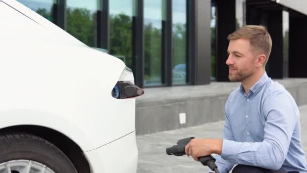 Hansome Tipo Sentado Cerca Nuevo Coche Eléctrico Moderno Sosteniendo Enchufe — Vídeo de stock