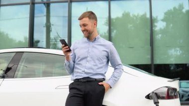 Portrait of young handsome european man, speaking on a video chat on his smartphone while leaning on his electric car, charging the battery at city power station.