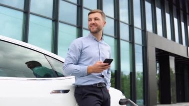 Aracın yanında duran ve cep telefonunu kullanan bir adamı ve elektrikli arabayı şarj ediyor..