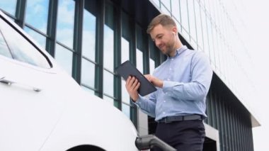 Young european businessman with wireless headphones, using tablet near electric car, charging the battery at city power station.