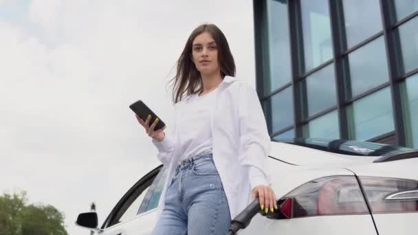 Młoda Kobieta Stoi Pobliżu Elektrycznego Samochodu Patrzy Smartfon Wypożyczony Samochód — Wideo stockowe